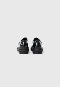 Zapatos de charol negros con puntera redondeada, suela de goma gruesa y diseño minimalista. Presentan una textura suave y un corte bajo en la parte trasera.