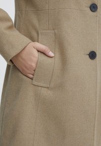 Beige wollen jas met een gladde textuur, voorzien van een zijzak, zwarte knopen en strakke lijnen in het stofontwerp.
