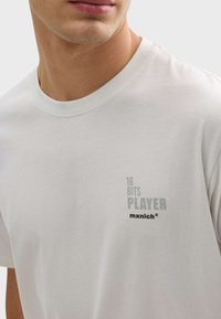 Svetlo šedé tričko s krátkymi rukávmi, okrúhlym výstrihom a nenápadným predným potlačou s nápisom "16 BITS PLAYER mxnich." Bavlnený materiál, hladká textúra.