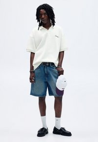 Polo bianco a maniche corte con logo ricamato, pantaloni corti in denim blu, cappellino bianco e viola, calze bianche alla caviglia e scarpe nere.