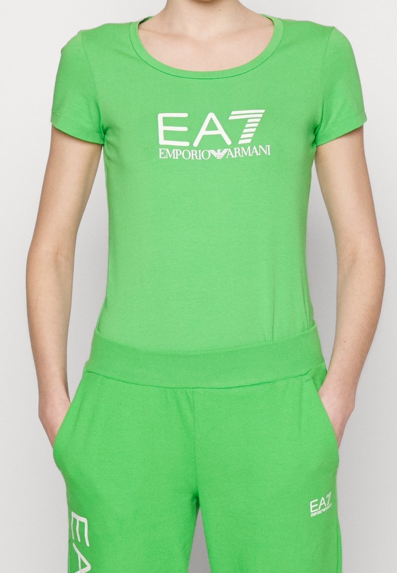 Zielony dwuczęściowy zestaw z bawełny, składający się z dopasowanego topu z krótkim rękawem i dekoltem w serek oraz dopasowanych spodni dresowych z elastycznym pasem. Wyraźnie widoczne logo EA7.