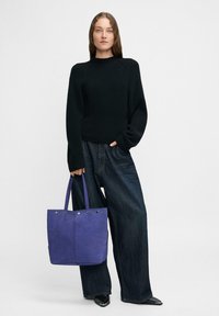 Kobieta stoi ubrana w czarny sweter, szerokie ciemne dżinsy, czarne spiczaste buty, trzymając dużą niebieską torbę typu tote z metalowymi oczkami.