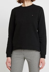 Sudadera negra con cuello redondo y puños acanalados. Presenta un pequeño logotipo bordado en el pecho. Combinada con pantalones a cuadros.