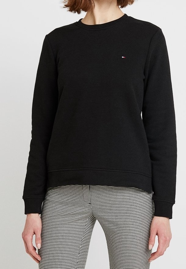 Sudadera negra con cuello redondo y puños acanalados. Presenta un pequeño logotipo bordado en el pecho. Combinada con pantalones a cuadros.