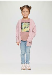 Helle rosa strukturierte Strickjacke mit Knöpfen, grafisches pinkes T-Shirt, hellblaue Jeans und pinke Sneakers mit weißen Akzenten. Mädchen mit zwei Duttfrisuren.