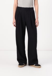 Pantalons larges noirs avec plis, fabriqués en tissu l�éger. Présente une taille ajustée et des poches latérales. Portés avec des baskets noires.