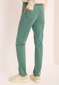 Pantalones de algodón verdes de corte ajustado, con bolsillos traseros y detalles de costura, combinados con zapatillas beige sobre un fondo claro.