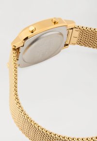 Relógio de pulso com pulseira em malha dourada visto de trás, com fundo da caixa em prateado e dois botões dourados de cada lado, sobre um fundo branco.