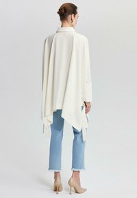 Châle long à manches drapées blanc sur une tenue en jean bleu. Le châle est doté d'un col et se noue sur les côtés. Des talons hauts beiges complètent le look.