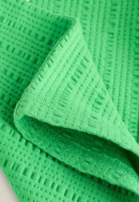 Tela verde texturizada con un patrón de cuadrícula, con bordes acanalados y una textura suave y tejida, doblada parcialmente sobre una superficie plana.