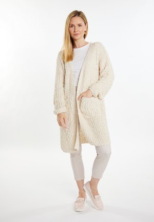 Cardigan cremoso con texture, maniche lunghe, apertura frontale e tasche, abbinato a pantaloni beige chiaro e scarpe slip-on rosa pallido.
