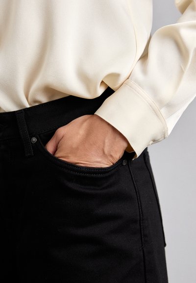 Black jeans with a smooth texture and classic design, featuring a front pocket, paired with a light-colored, silky blouse.