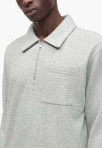 Gros plan d'une personne portant une chemise gris clair à manches longues avec un col zippé et une poche poitrine.
