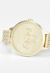 Montre analogique ton or avec cadran texturé, trois sous-cadrans pour le jour, la date et le format 24 heures, et un bracelet en métal relié.
