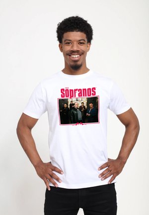 Smilende mand iført en hvid T-shirt med et rødt "The Sopranos"-logo og et gruppefoto af karakterer fra tv-showet.