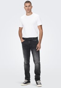 Herenoutfit met een getailleerd wit T-shirt en donkergrijze jeans. De jeans hebben een lichte vervaging, met een zwarte riem en zwarte sneakers.