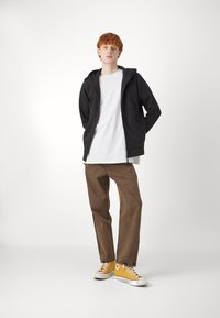 Felpa nera con zip, t-shirt bianca, pantaloni dritti marroni e sneaker in tela gialla con suole bianche. Design semplice, look casual.