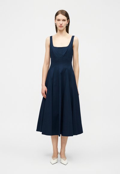 Donna che indossa un abito midi blu navy senza maniche con scollo quadrato e tacchi bianchi, in piedi contro uno sfondo chiaro e uniforme.