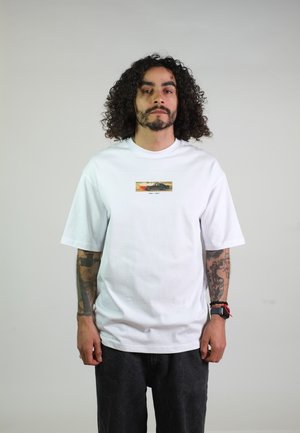 Man met krullend haar en tatoeages die een wit T-shirt draagt met een kleine autografiek, staand tegen een effen lichte achtergrond.