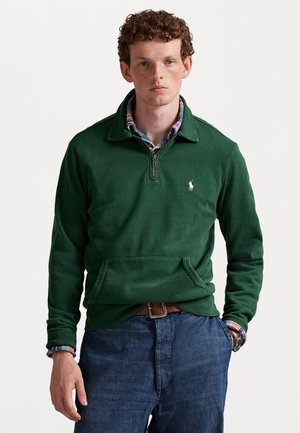 Jonge man met krullend rood haar, gekleed in een groene kwart-zip trui over een geruit shirt en blauwe spijkerbroek, staand tegen een effen achtergrond.
