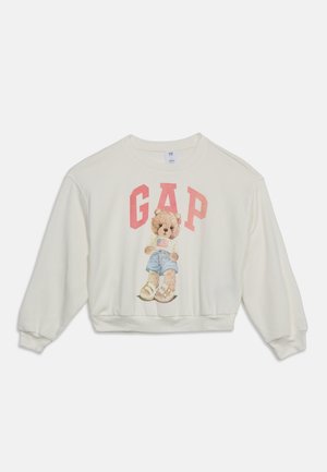 Witte trui met een teddybeer die korte broek en sandalen draagt, een Amerikaanse vlag vasthoudend, met grote roze "GAP"-tekst erboven.