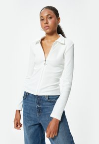 Maglia bianca a coste con zipper e colletto, con maniche lunghe e una vestibilità aderente. Abbinata a jeans blue a vita alta. Dettaglio della zip in argento.