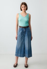 Top in cotone verde menta a costine, culotte in denim blu a vita alta e gamba larga con una cintura marrone e scarpe basse intrecciate scure. Sfond semplice.