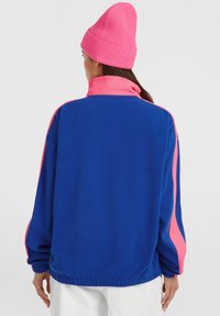 Blå fleecejakke med pink detaljer på kraven og ærmerne. Ribbet pink hue. Løs pasform med elastiske manchetter og linning.