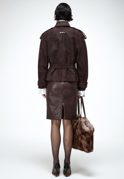 Nő sötétbarna velúr dzsekiben, bőr szoknyában, fekete áttetsző harisnyában, fekete magassarkúban, barna műszőrme kézitáskát tartva, háttal állva.