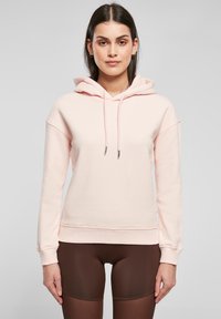 Lys rosa hettegenser laget av myk stoff, med snor, oversized passform og ribbede mansjetter. Kombinert med brune leggings.