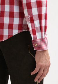 Camicia a quadri rossi e bianchi con maniche lunghe, dotata di polsino con bottone e motivo a quadri in contrasto sul polsino, indossata su un tessuto marrone scuro.