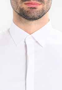 Biała koszula na guziki z klasycznym kołnierzykiem, gładką fakturą i pojedynczym guzikiem przy szyi, widoczne przeszycia na listwie guzikowej.