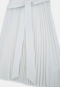 Biała plisowana spódnica z szerokim pasem i kokardą, z pionowymi plisami ciągnącymi się od pasa do dołu.