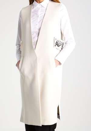 Femme portant une longue veste crème sans manches avec des boutons décoratifs sur une chemise blanche boutonnée et un pantalon noir, les mains dans les poches.