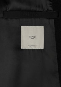 Intérieur en tissu noir avec une étiquette carrée blanche portant l'inscription "MNG MAN", taille moyenne, fabriqué au Vietnam, avec une étiquette noire au-dessus cousue "MNG MAN".