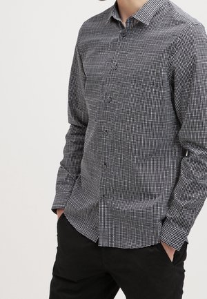 Homme portant une chemise à manches longues à carreaux noirs et blancs, les mains dans les poches, associée à un pantalon noir sur un fond uni.