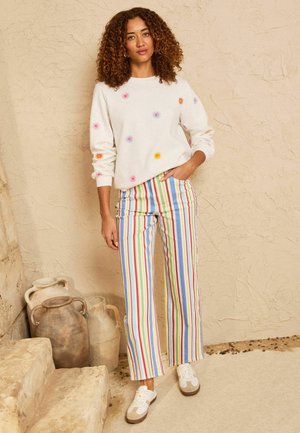 Frau mit lockigem Haar steht an einer strukturierten Wand, trägt ein weißes Sweatshirt mit bunten Sonnenmustern, Regenbogen-gestreifte Hosen und weiße Turnschuhe.