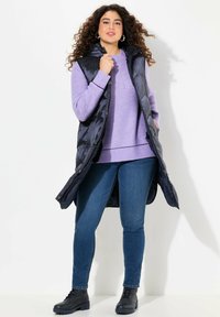 Lång svart pufferväst över en lavendeltröja, kombinerat med blå jeans och svarta ankelboots. Glansig textur och huva.