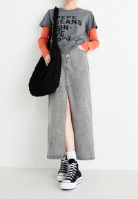 Falda maxi de denim gris con una hendidura frontal, combinada con una camiseta gris que presenta texto negro y una camiseta de manga larga naranja debajo. Zapatillas negras.