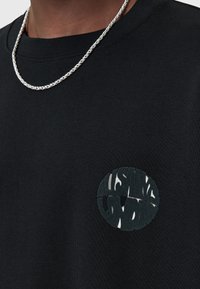 Sudadera de algodón negra con un parche redondo que lleva el texto en verde y blanco "ALWAYS LDN". Cuello redondo con canalé y tejido texturizado. Collar de cadena plateado visible.