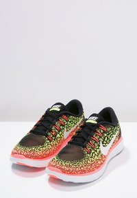 Černé a neonově žluté síťované běžecké boty s růžovou podrážkou, které mají bílý Nike swoosh a šněrovací design. Vzorovaná textura po celé ploše.