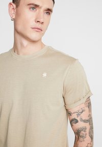 Beige T-shirt met korte mouwen en een ronde ronde halslijn en opgerolde mouwen. Heeft een klein, subtiel logo op de borst. Zachte, gladde stof.