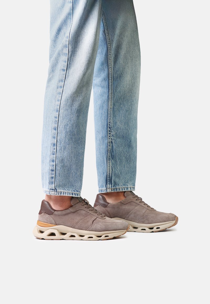Personne portant un jean bleu clair et des baskets en daim gris avec des semelles couleur crème sur fond blanc.