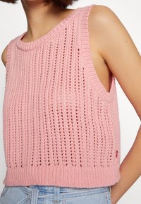 Pull sans manches en tricot rose avec une texture côtelée, des côtés ouverts et une coupe courte, avec une petite étiquette sur l'ourlet.
