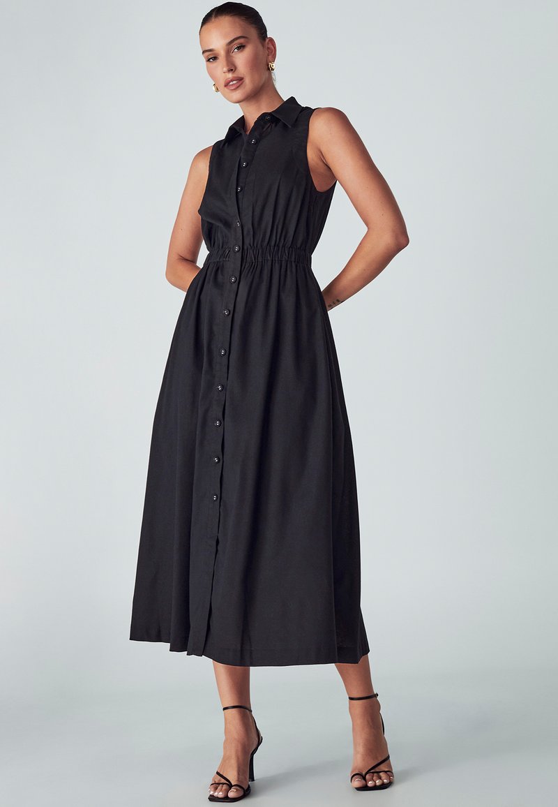 Juoda be rankovių suknelė, pasižyminti mygtukų užsegimu, elastine juosmens dalimi ir laisvai krentančia sijone, siekiančia vidurinę blauzdą. Lygus audinio paviršius.