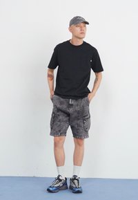 T-shirt noir avec poche, shorts cargo gris à motif, chaussettes blanches et chaussures de sport bleues. Une casquette grise complète la tenue décontractée.