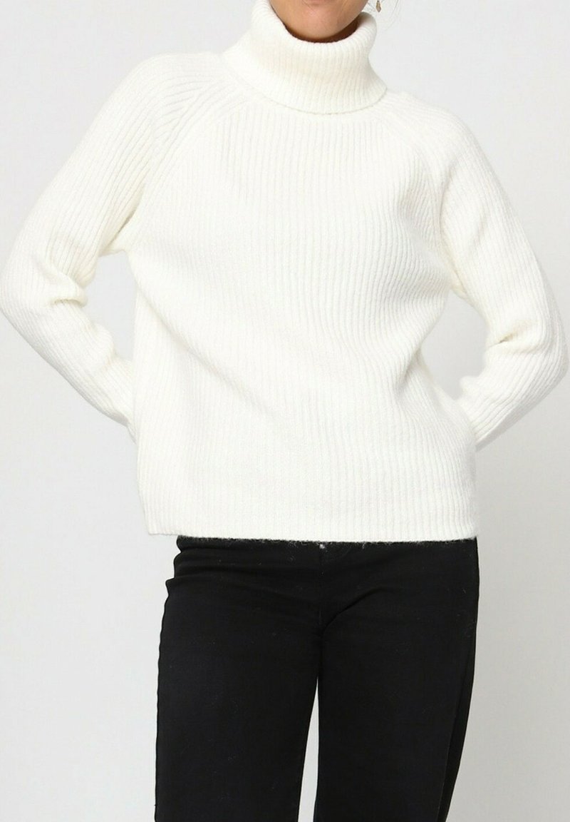 Pull à col roulé blanc côtelé avec une coupe décontractée, présentant des manches longues et un ourlet droit. Associé à un pantalon noir.