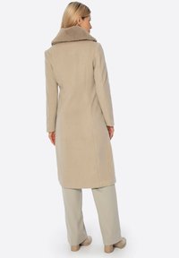 Beżowy wełniany płaszcz z dużym, miękkim kołnierzem, dopasowany fason i długość do połowy uda. W zestawie z jasnobeżowymi spodniami i prostymi butami.