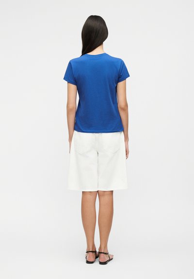 Γυναίκα που στέκεται με την πλάτη γυρισμένη, φοράει μπλε μπλουζάκι με κοντά μανίκια, λευκά σορτς μέχρι το γόνατο και μαύρα πέδιλα με ανοιχτά δάχτυλα σε λευκό φόντο.