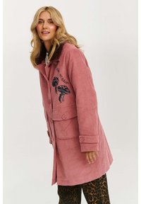 Ružový kordurový kabát s hnedým kožušinovým golierom, veľkými gombíkmi a vyšívaným dizajnom hríbu; obsahuje dve predné vrecká a má strednú dĺžku.